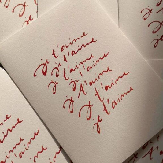 "Carte Je t'aime répétée en rouge, posée sur plusieurs autres cartes similaires, avec un design manuscrit élégant"