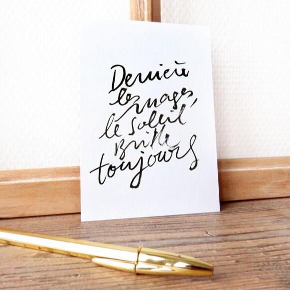 "Carte Derrière les nuages, le soleil brille toujours avec une écriture manuscrite noire, posée sur une table en bois à côté d'un stylo doré"