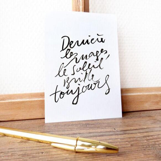 "Carte Derrière les nuages, le soleil brille toujours avec une écriture manuscrite noire, posée sur une table en bois à côté d'un stylo doré"