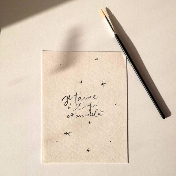"Carte Je t'aime à l'infini et au-delà, avec des petites étoiles noires et un pinceau noir, posée sur un fond blanc"
