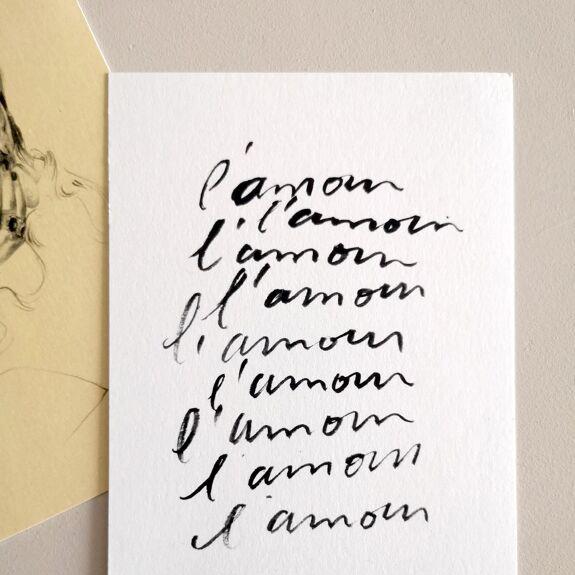 "Carte postale avec répétition manuscrite du mot L'amour, en noir sur papier blanc, posée à côté d'une illustration"