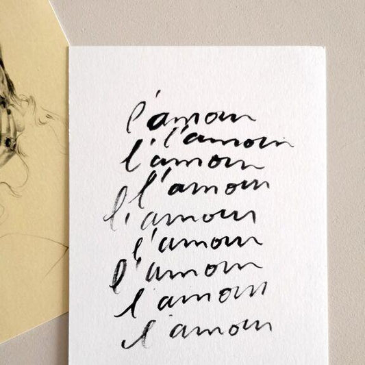 "Carte postale avec répétition manuscrite du mot L'amour, en noir sur papier blanc, posée à côté d'une illustration"